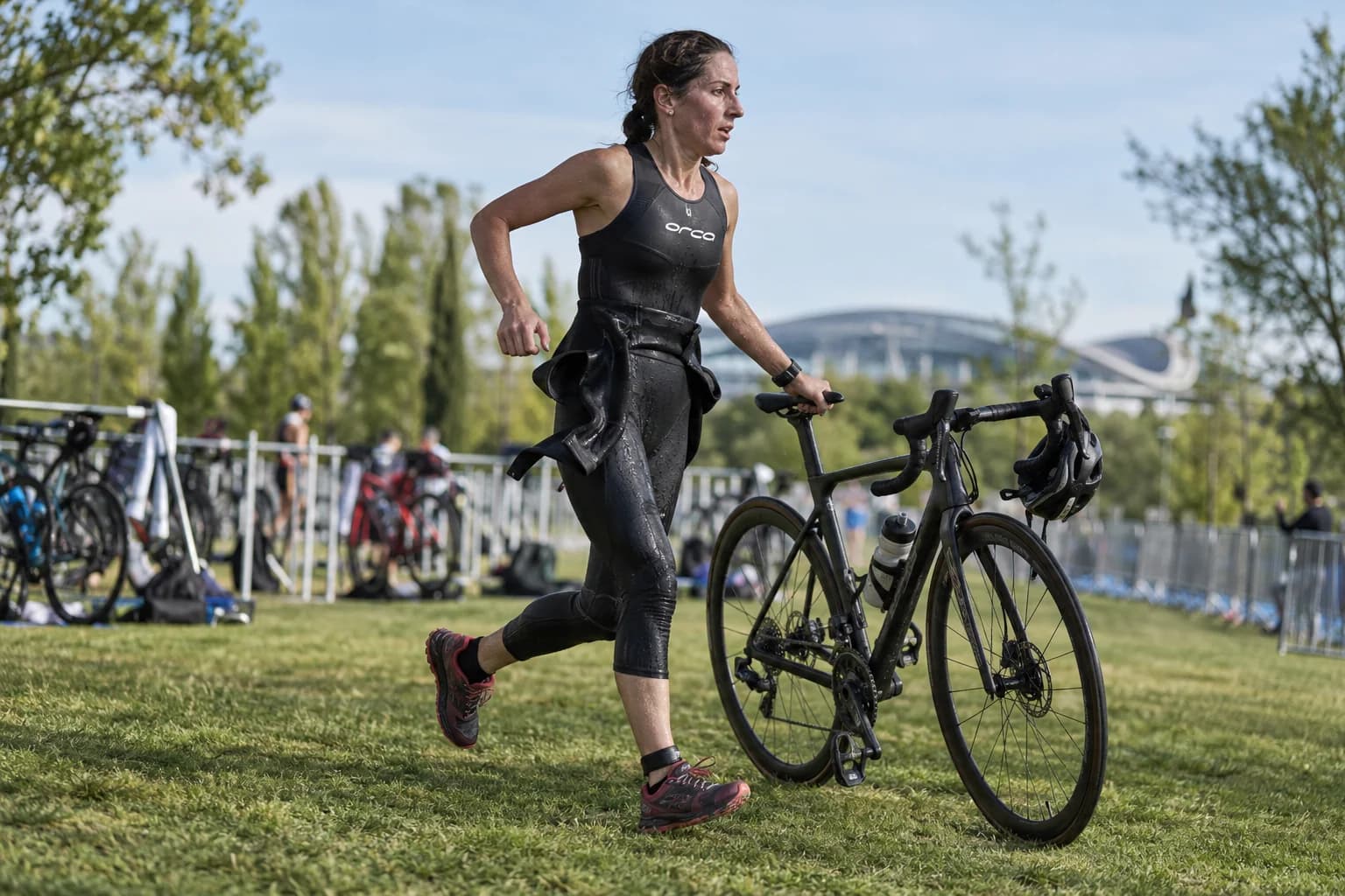 AI-gegenereerd sfeerbeeld: triatlete die met haar fiets door een wisselzone in een park rent